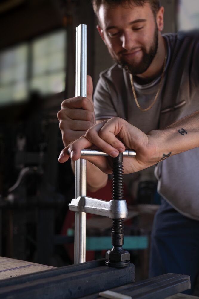 Bearded man tightens a heavy-duty F-Style clamp on metal bars in a workshop.