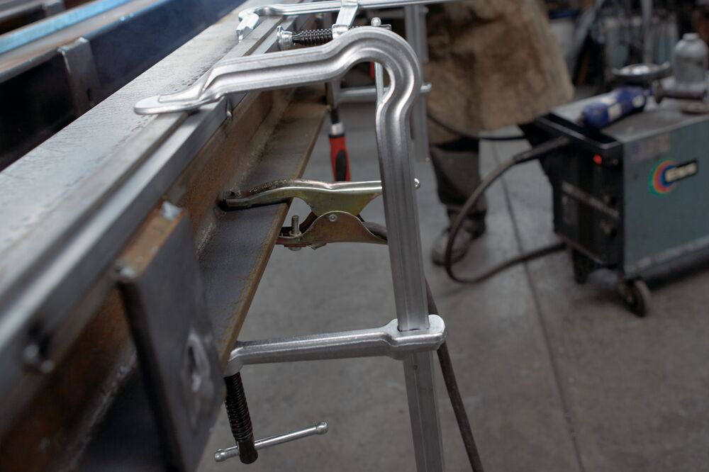 C clamp securing a metal bar to a workbench in a busy workshop with a welding machine in the background
