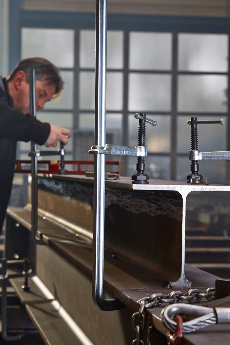 Heavy duty all steel F-style welding clamp grips a metal channel on a workbench with a technician in the background