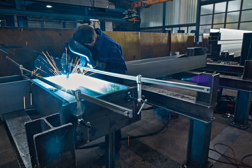 Welder at work with a steel beam secured by a large clamp system in an industrial workshop.