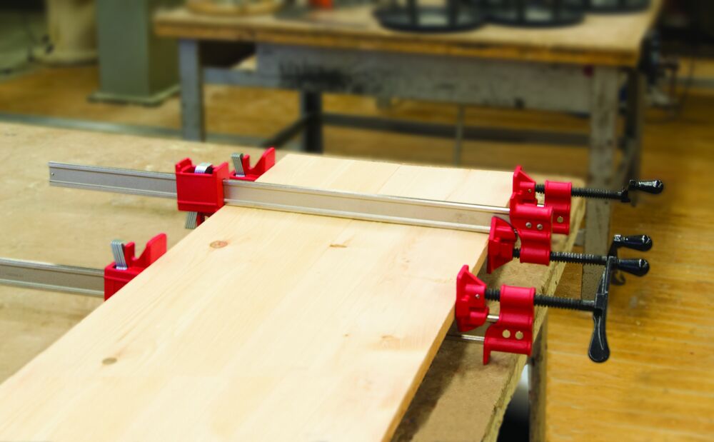 Multiple red Bessey I-Beam Bar Clamps along a silver rail securing a wooden board on a workbench
