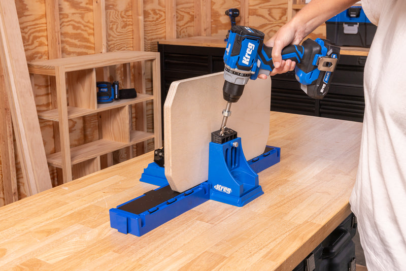 Blue Kreg K4 pocket-hole jig workstation mounted on a benchtop with a cordless drill drilling into a plywood panel.