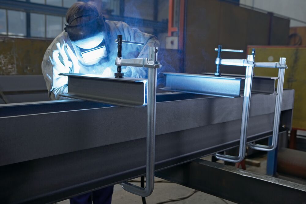 Worker in protective gear uses a heavy-duty F-Style welding clamp to hold a steel beam.