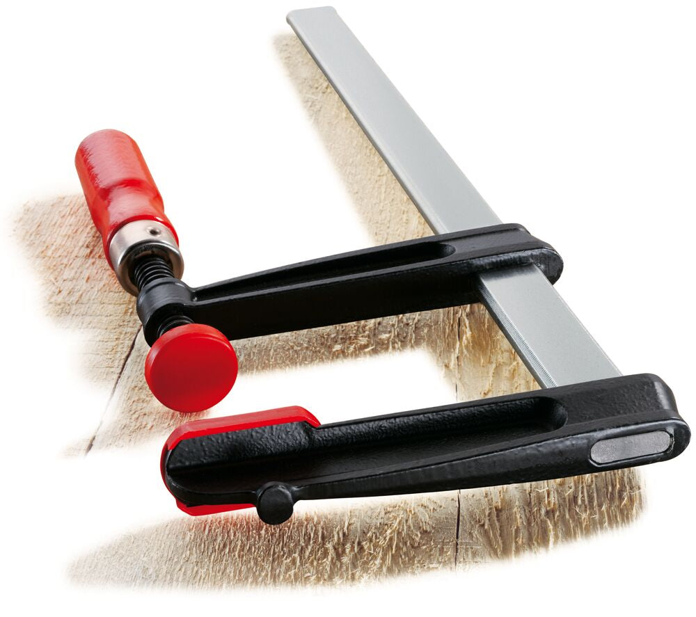 Bessey flat rail clamp in use grips a wood piece to a workbench with a red adjustment knob visible