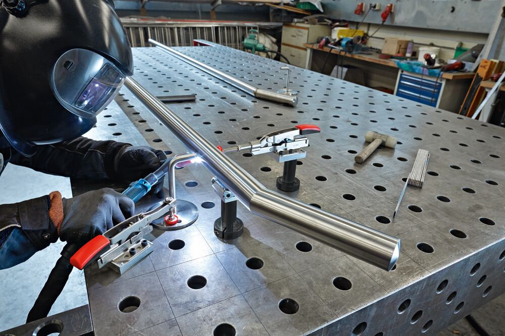 Welding operator secures a long metal tube with a Bessey toggle clamp mounted on a perforated welding table.